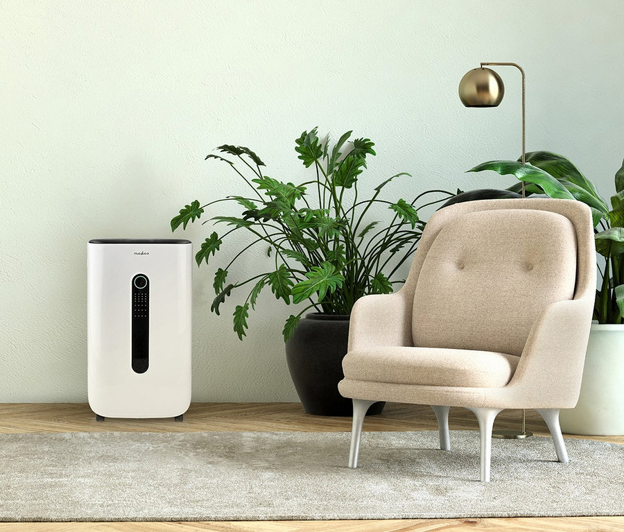 Ein weißer Nedis Smart Luftentfeuchter steht auf einem Holzboden neben einer großen Grünpflanze, einem beigen Sessel und einer goldenen Stehlampe auf einem hellgrauen Teppich vor einer hellgrünen Wand.