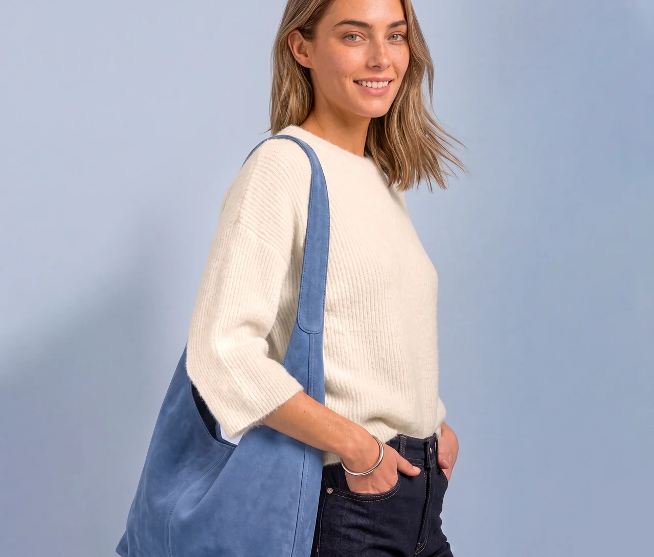 Eine lächelnde Frau mit blonden Haaren trägt einen cremefarbenen Street One Strickpullover, dunkle Jeans und eine hellblaue Umhängetasche. Ihre rechte Hand steckt in der Hosentasche.
