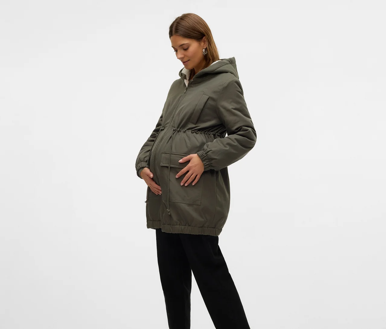 Schwangere Frau in einer grünen Kapuzenjacke und schwarzen Hosen, die nach unten schaut und ihren Bauch mit beiden Händen hält.