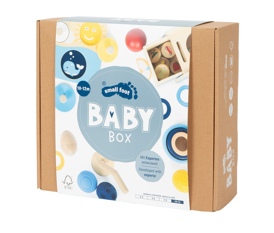 Eine braune Kartonbox mit einem hellblauen Etikett, auf dem "BABY BOX" und "small foot" steht. Das Etikett zeigt Illustrationen von Holzspielzeug wie Ringen, einer Sonne, einem Wal und einer Sortierbox. Altersangabe 10-12m.