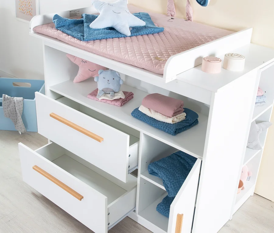 Weißer Wickeltisch mit rosa Wickelauflage, blauer Decke und Sternkissen oben. Er hat offene Ablagen mit einem blauen Stofftier und gefalteter Kleidung, zwei teilweise geöffnete Schubladen mit Holzgriffen und einen Seitenschrank.