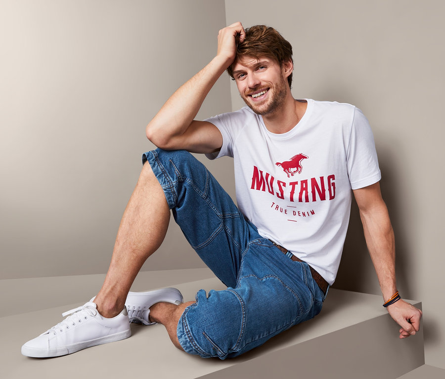 Mann sitzt und trägt Jeans-Bermuda-Shorts »Mustang« und ein T-Shirt.