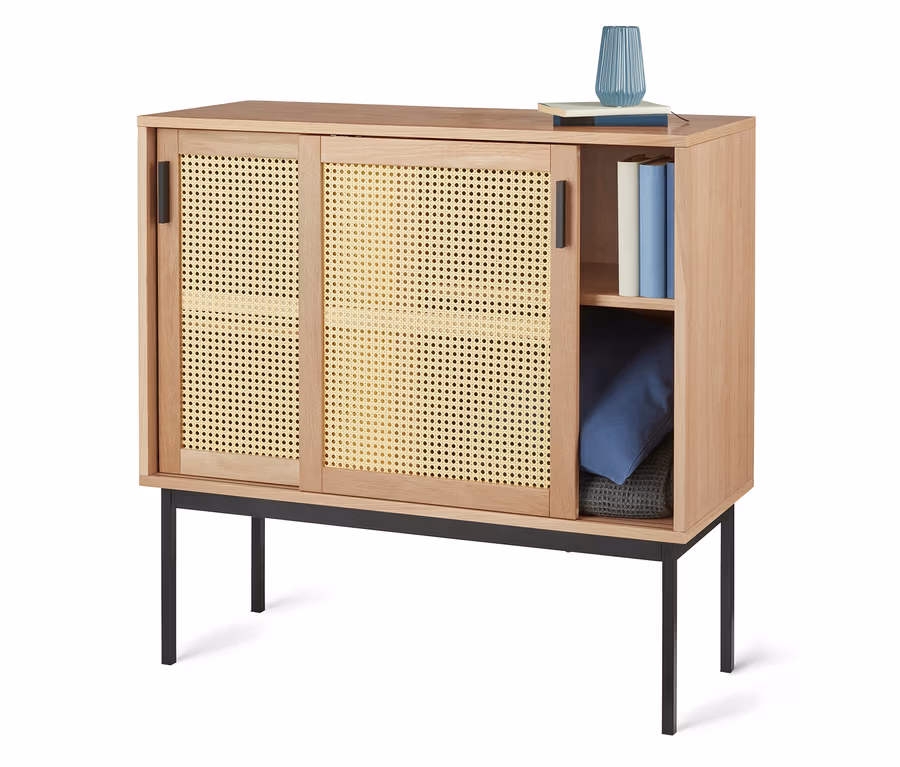 Sideboard mit geflochtenen Türen auf schwarzen Füßen. Oben Vase und Bücher, innen Regal mit Büchern und Kissen.