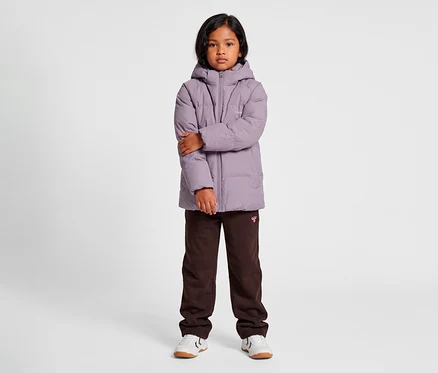 Ein junges Kind mit dunklen Haaren steht frontal da und trägt eine mauvefarbene hummel® »HMLJR All Weather Puffer Jacket« mit Kapuze, dunkelbraune Jogginghosen und weiße Turnschuhe. Die Arme des Kindes sind verschränkt.