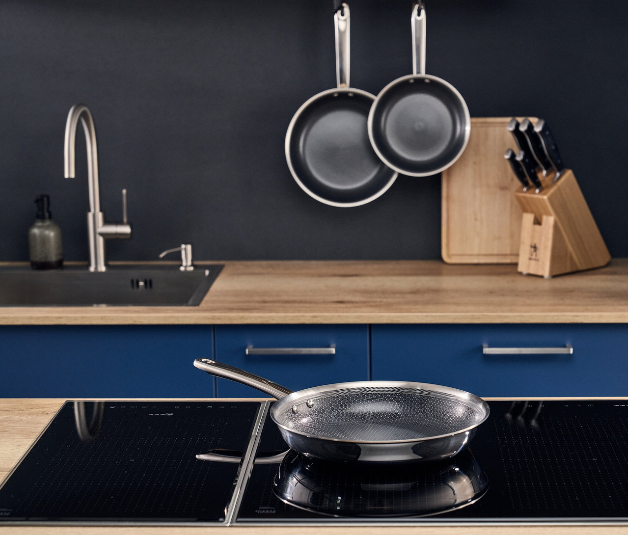 Moderne Küche mit dunkelblauer Wand und Holzarbeitsplatte. Eine Henckels by ZWILLING »Valley« Bratpfanne aus einem 3-teiligen Set steht auf einem schwarzen Induktionskochfeld. Zwei weitere Bratpfannen hängen an der Wand, neben einem Messerblock und Schneidebrett.