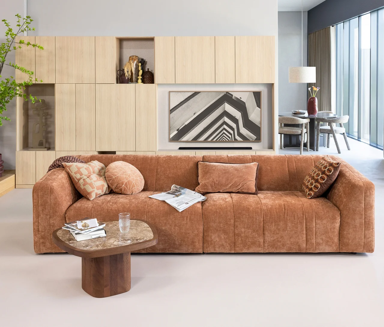 Ein modernes Wohnzimmer mit einem großen rostfarbenen Sofa, einem Couchtisch und einer hölzernen Schrankwand mit Fernseher. Auf dem Sofa liegen vier WOOOD Dekokissen »Luvo Velvet« in verschiedenen Formen und Mustern, darunter ein rechteckiges rostfarbenes Samtkissen, ein rundes pfirsichfarbenes Kissen und zwei quadratische Kissen mit Mustern. Im Hintergrund ist ein Essbereich zu sehen.
