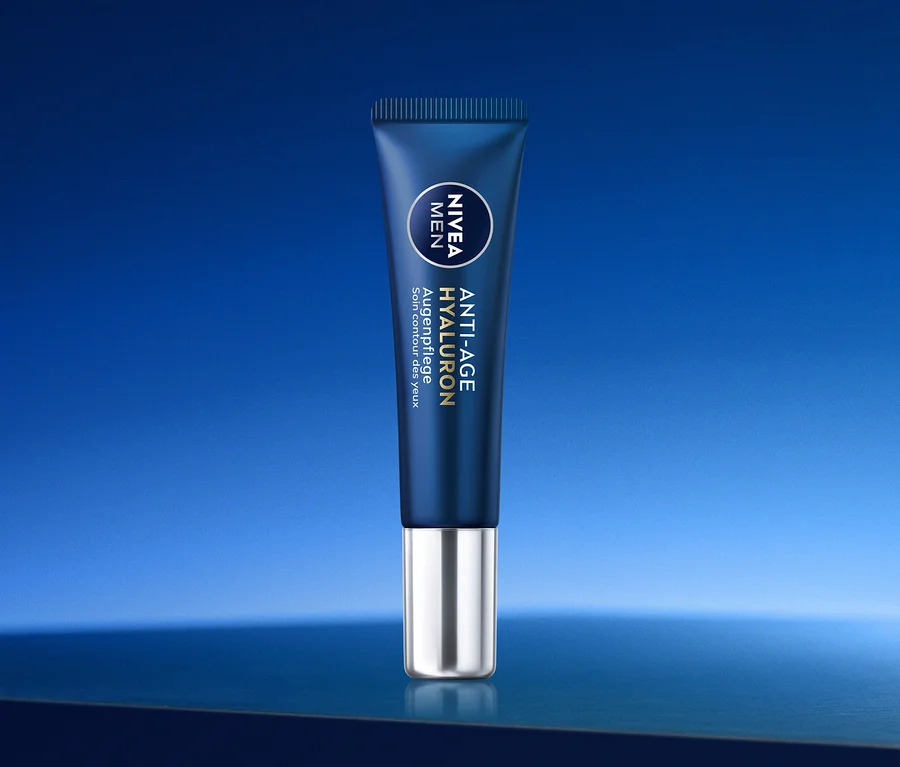 Dunkelblaue Tube Nivea Men Anti-Age Hyaluron Augenpflege mit silbernem Verschluss/Applikator, schräg auf einer reflektierenden blauen Oberfläche vor blauem Farbverlaufshintergrund stehend.