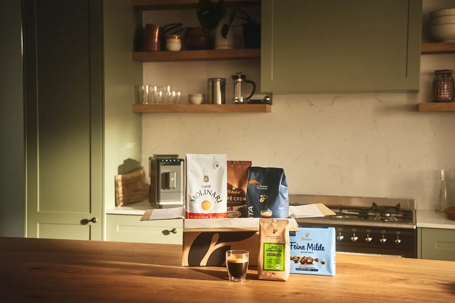 Auf einem Holztisch stehen ein Glas Espresso und eine offene Kiste mit Molinari- und Tchibo-Kaffeebohnen.