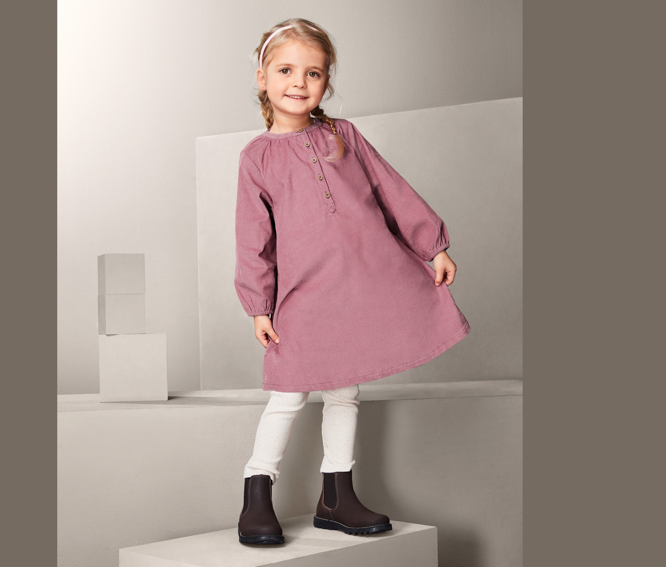 Mädchen steht auf einem weißen Würfel und trägt ein Cordkleid, weiße Leggings und braune Schuhe.