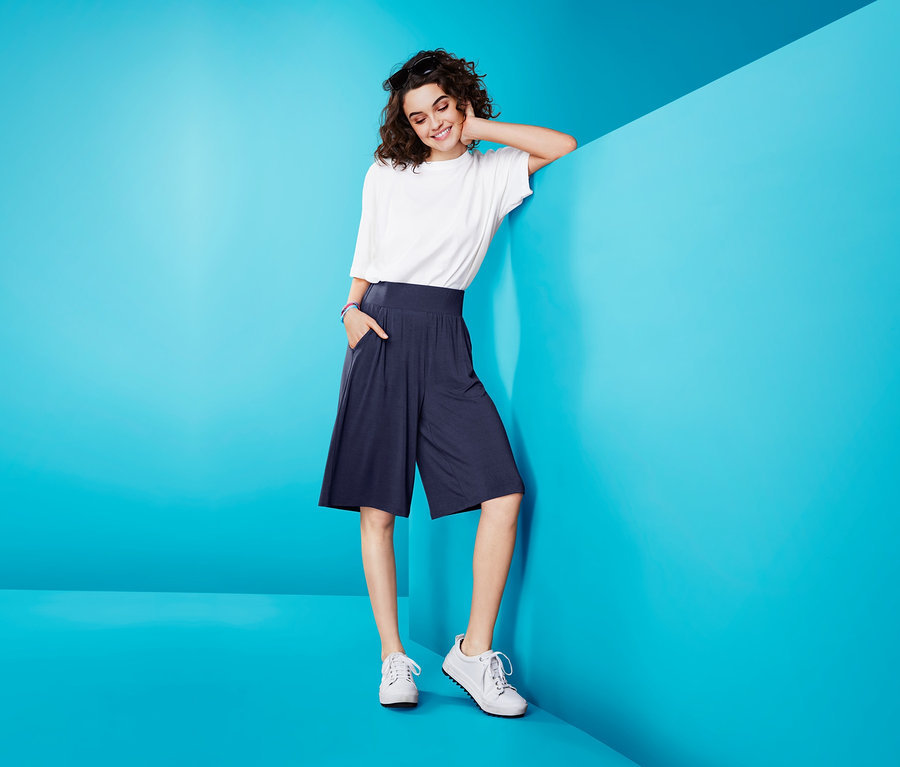 Frau lehnt an blauer Wand, trägt weißes T-Shirt und Hosenrock.