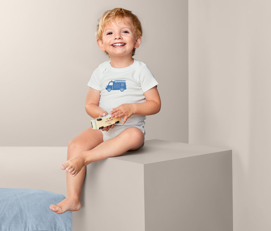 Ein lächelnder Säugling sitzt und hält ein hölzernes Spielzeugauto. Er trägt einen weißen Kurzarmbody mit einem blauen Feuerwehrauto-Aufdruck. 3 Kurzarm-Bodys.