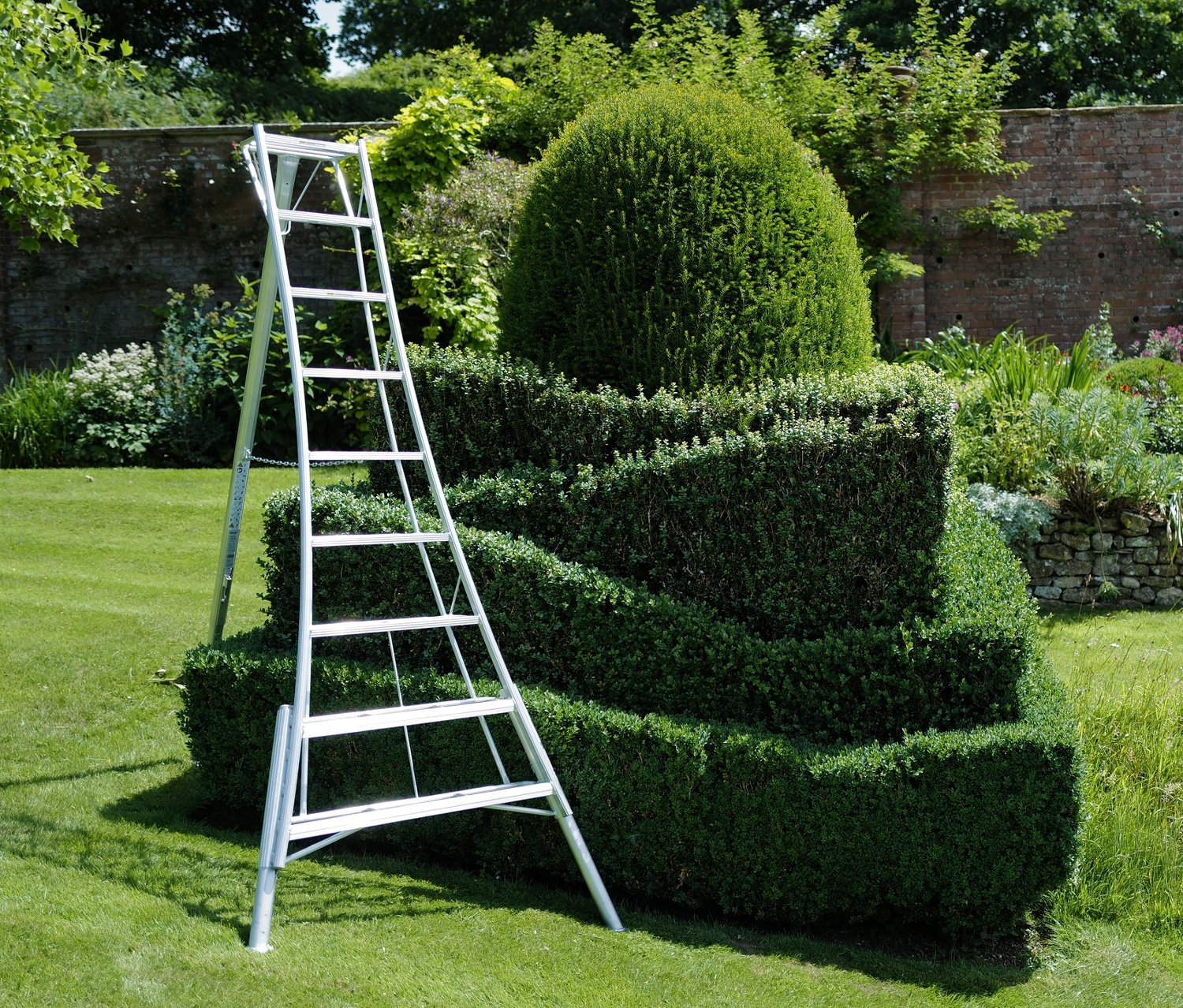 Eine silberne Leiter steht neben einem grünen Busch in einem Garten. Im Hintergrund ist der Niwaki-3-Holm-Gartenleiter »GSCS-270« zu sehen.