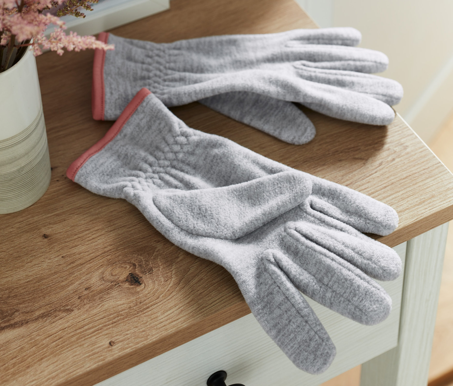 Zwei graue Fleece-Handschuhe liegen auf einem Holztisch neben einer Vase mit rosa Blumen.