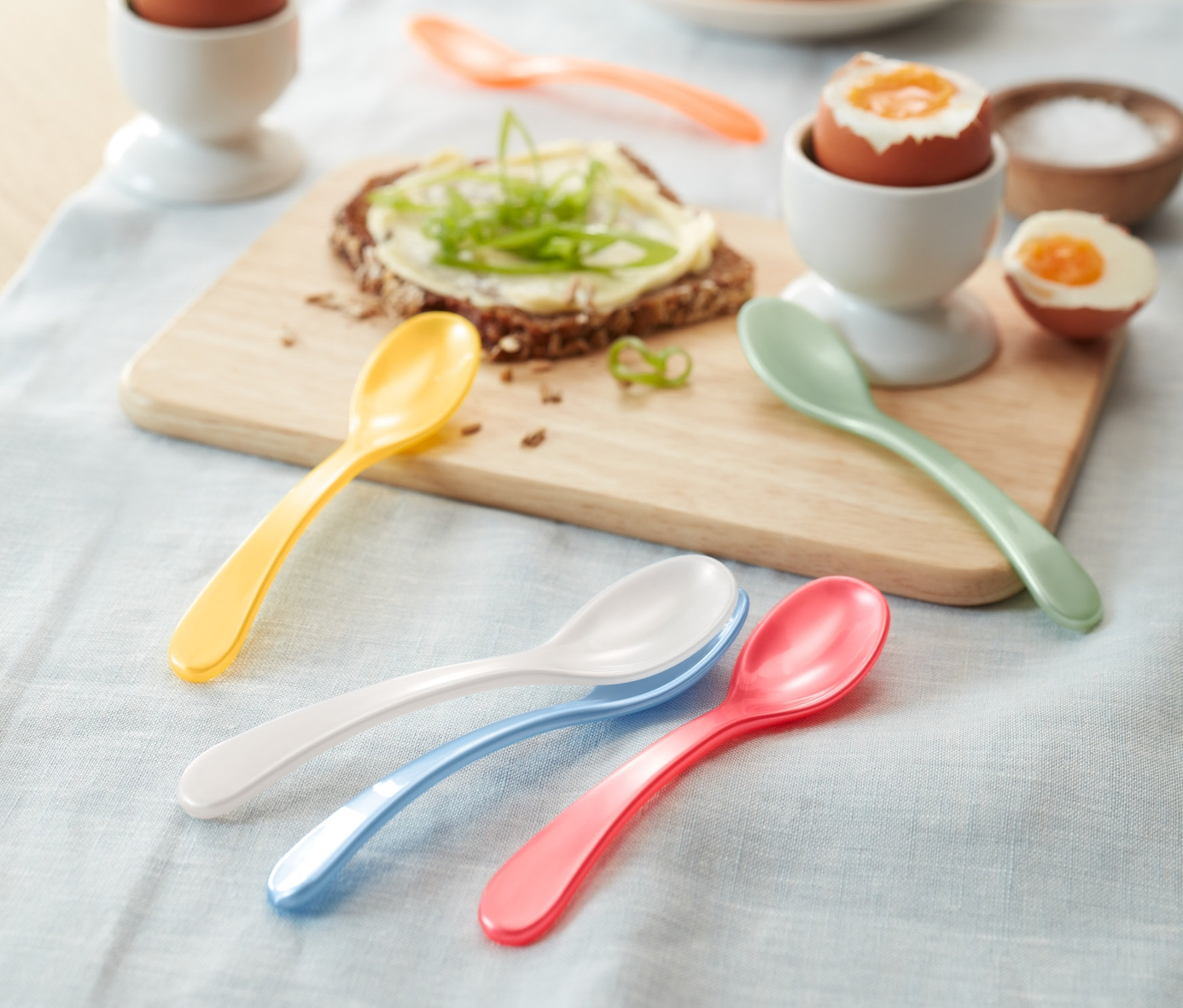 Bunte Eierlöffel, Eier im Eierbecher und ein Butterbrot liegen auf dem Tisch.