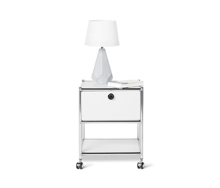 Auf einem weißen Hintergrund steht ein weißer Beistelltisch Metall »CN3« mit Schublade, auf dem eine Lampe steht und ein Buch liegt.