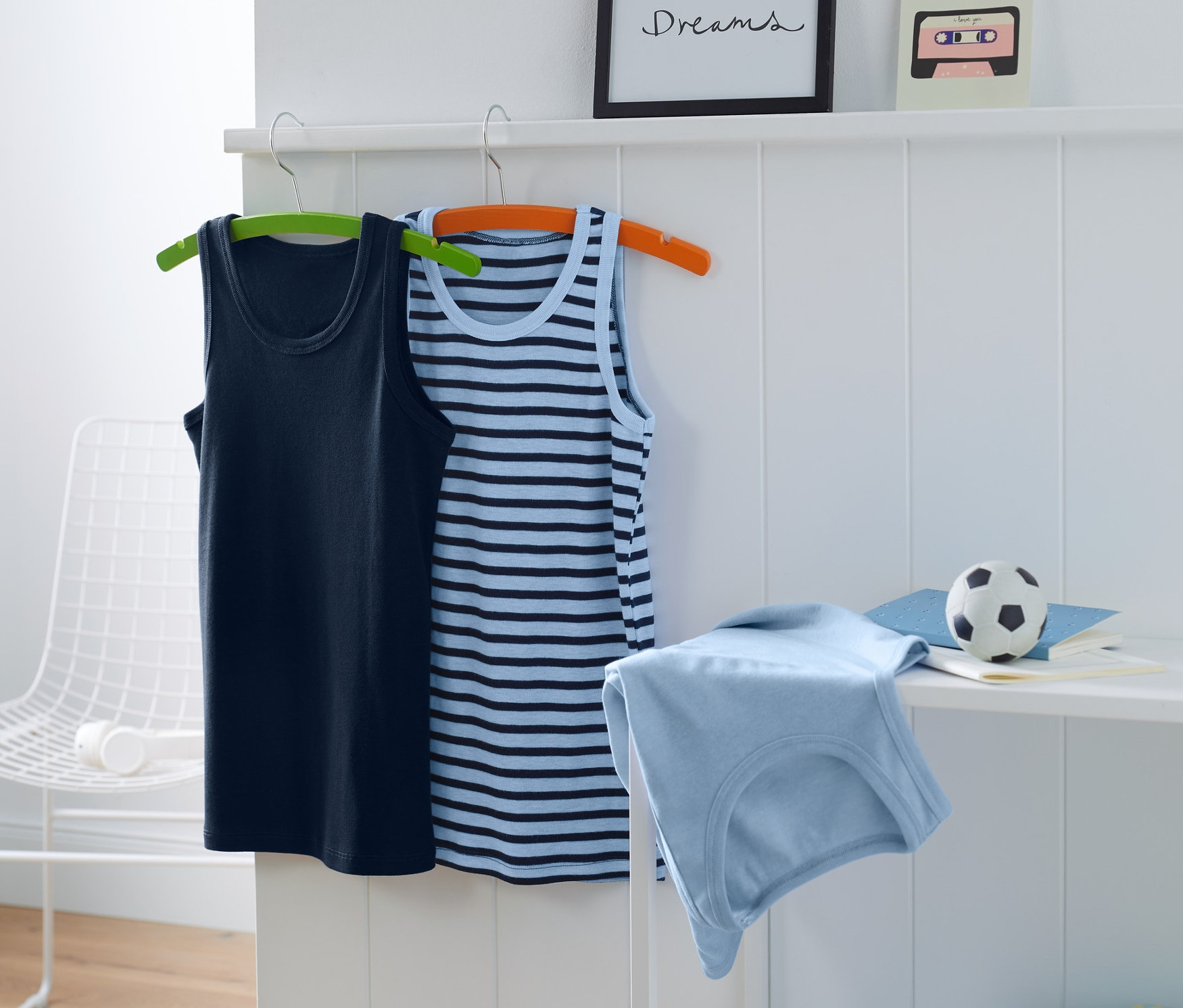 Zwei blaue Jungen-Unterhemden hängen an Kleiderbügeln, eines uni blau, eines blau gestreift, ein drittes blaues Unterhemd liegt auf einem Regal.
