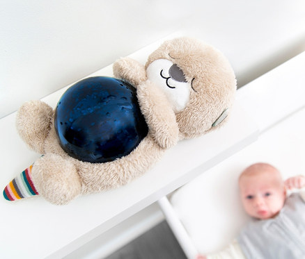 Ein ZAZU-Ozeanprojektor »Otto« Stofftier liegt auf einem weißen Regal. Im Hintergrund liegt ein Säugling in einem Kinderbett.