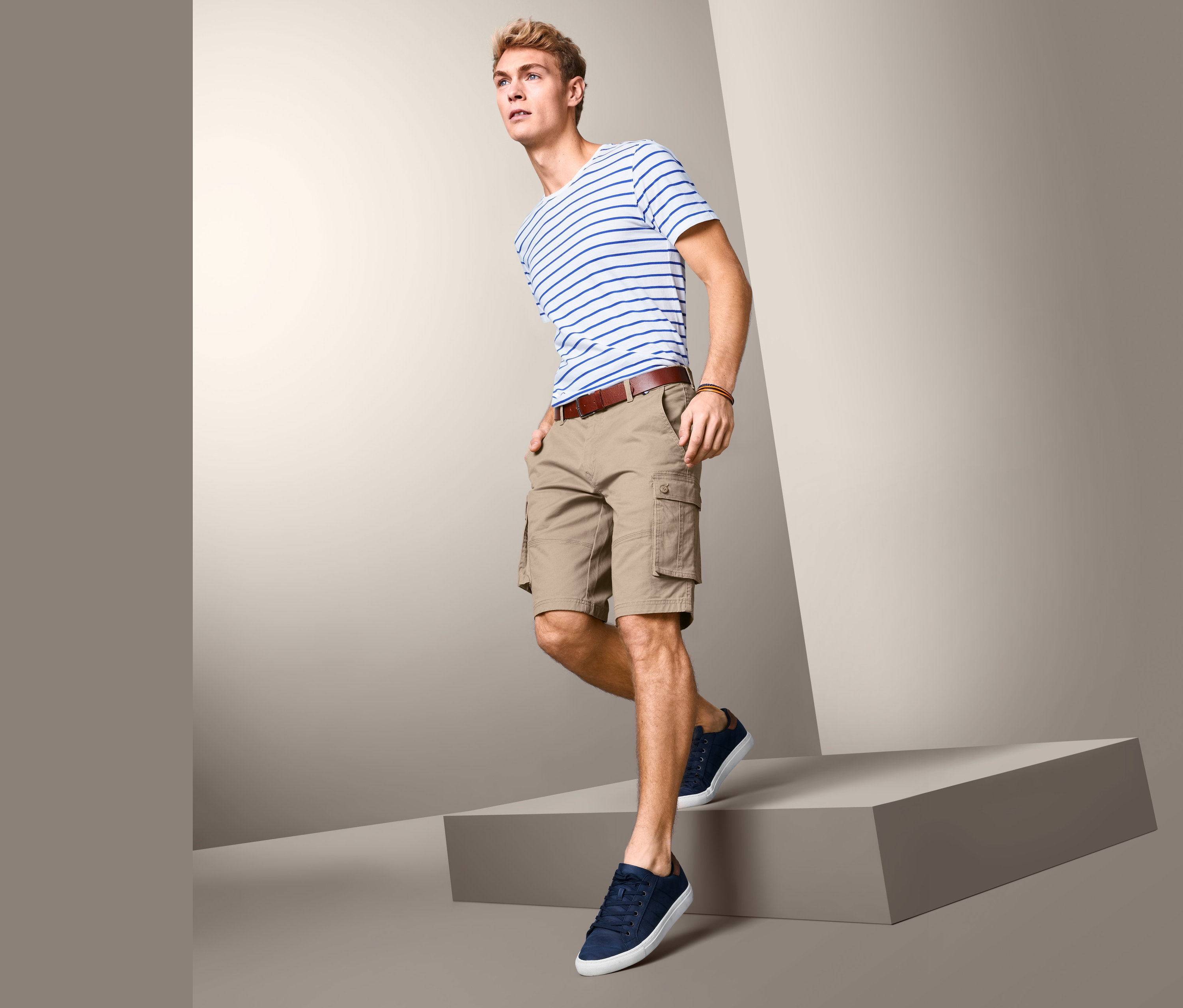 Mann posiert auf einer grauen Plattform in einem gestreiften T-Shirt und beigen Cargo-Bermudas.