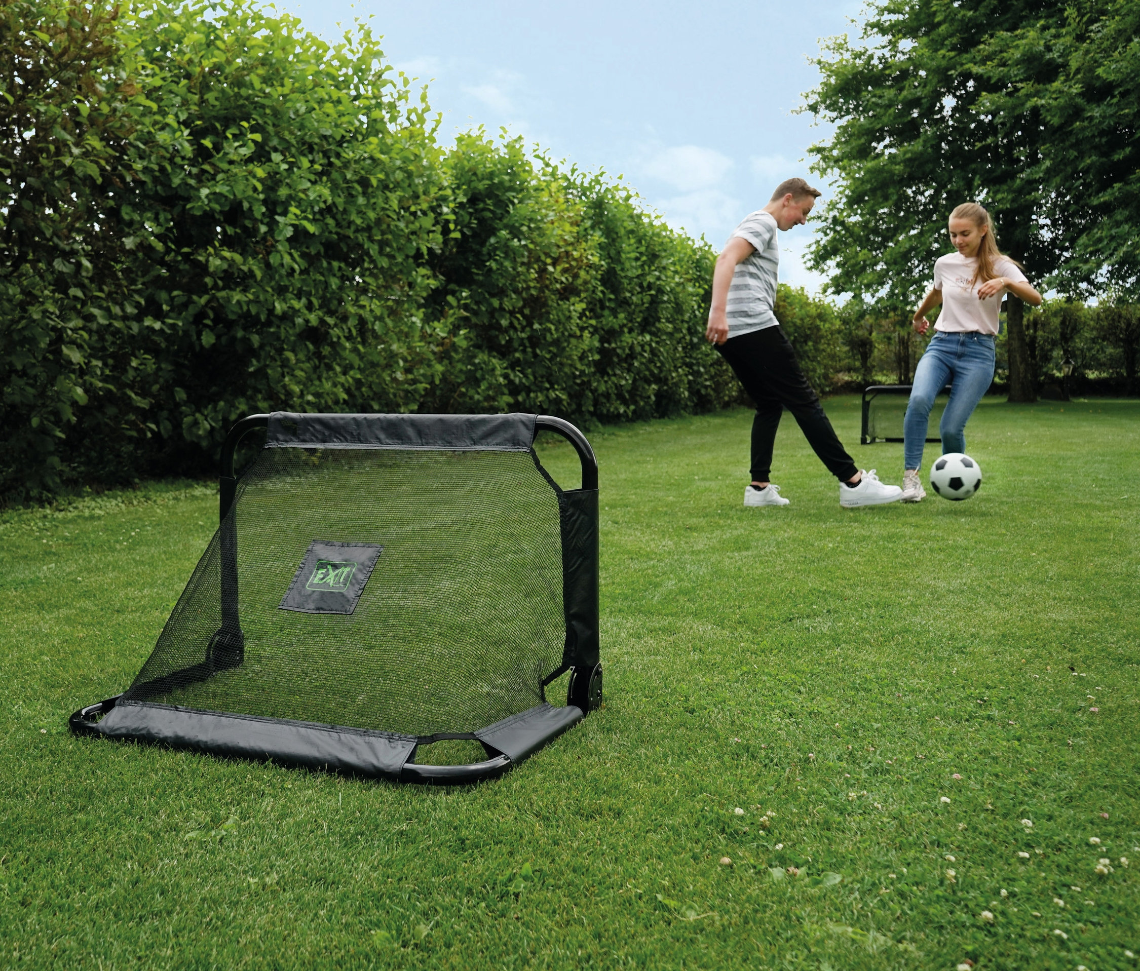 Zwei Personen spielen Fußball in der Nähe von 2 EXIT Mini-Fußballtoren »Pico« auf einer Rasenfläche.