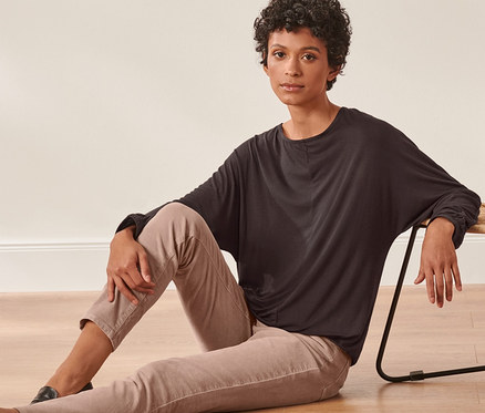 Frau sitzt neben Korbstuhl auf Boden, trägt Fledermausshirt.