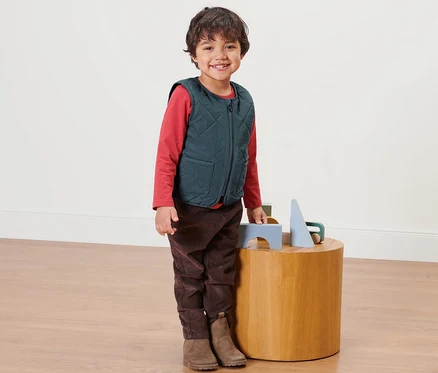 Ein Junge steht neben einem Holztisch und trägt eine Kinder-Wendesteppweste.