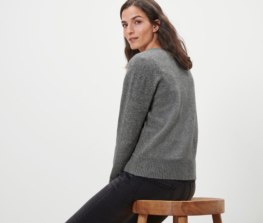 Frau sitzt auf einem Holzhocker und trägt eine grau melierte Strickjacke mit Wolle.