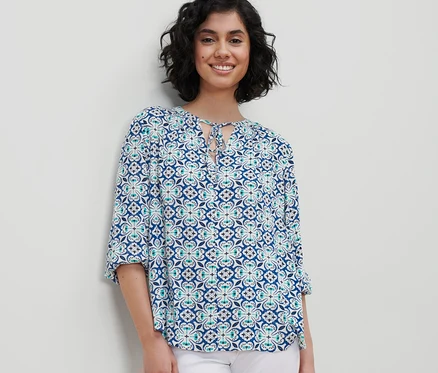 Frau trägt eine Tunika-Bluse mit Alloverprint und weiße Hose.