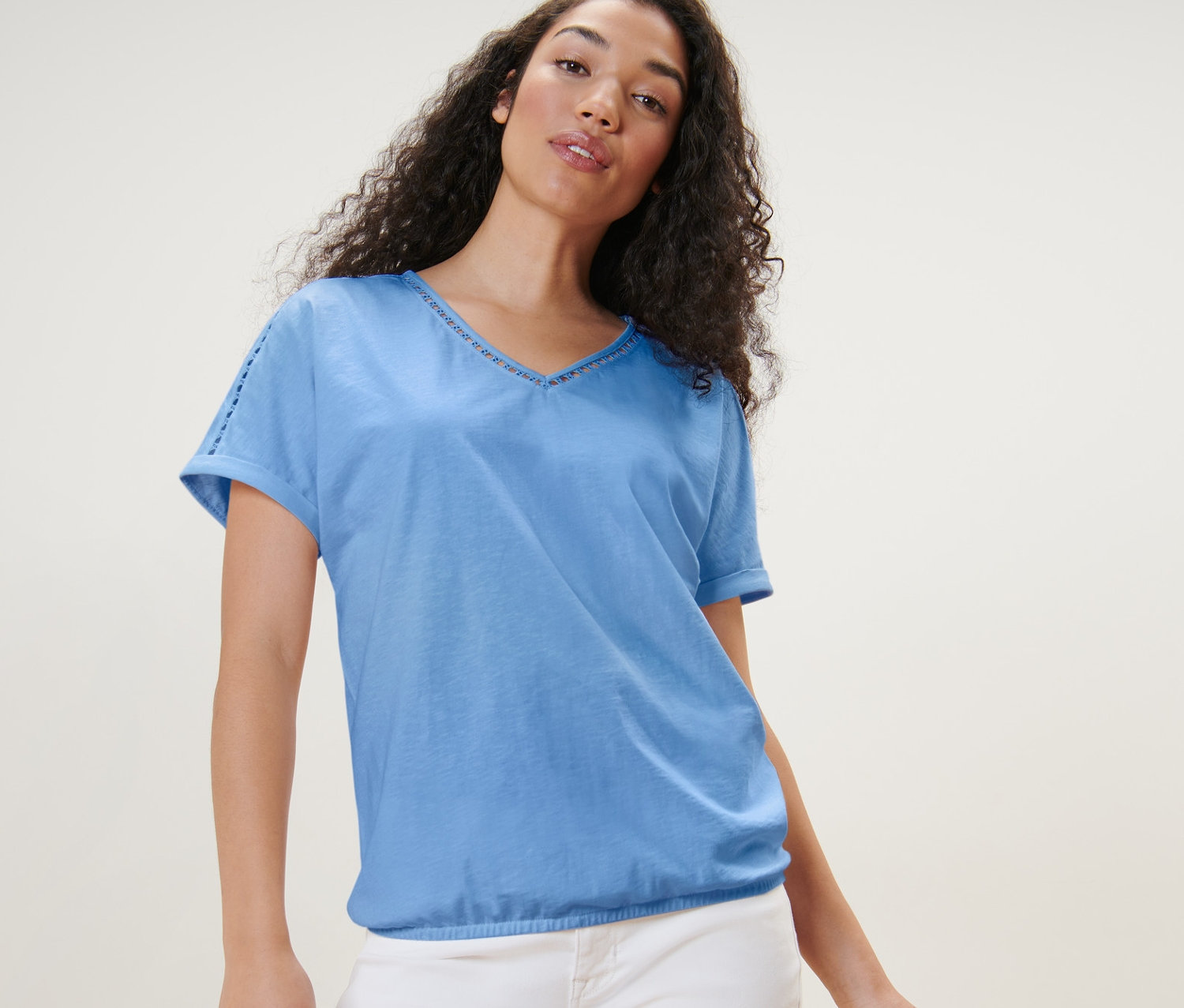 Frau posiert in hellblauem Blusenshirt mit Crochet-Tape und weißen Hosen neben einem Holzhocker.