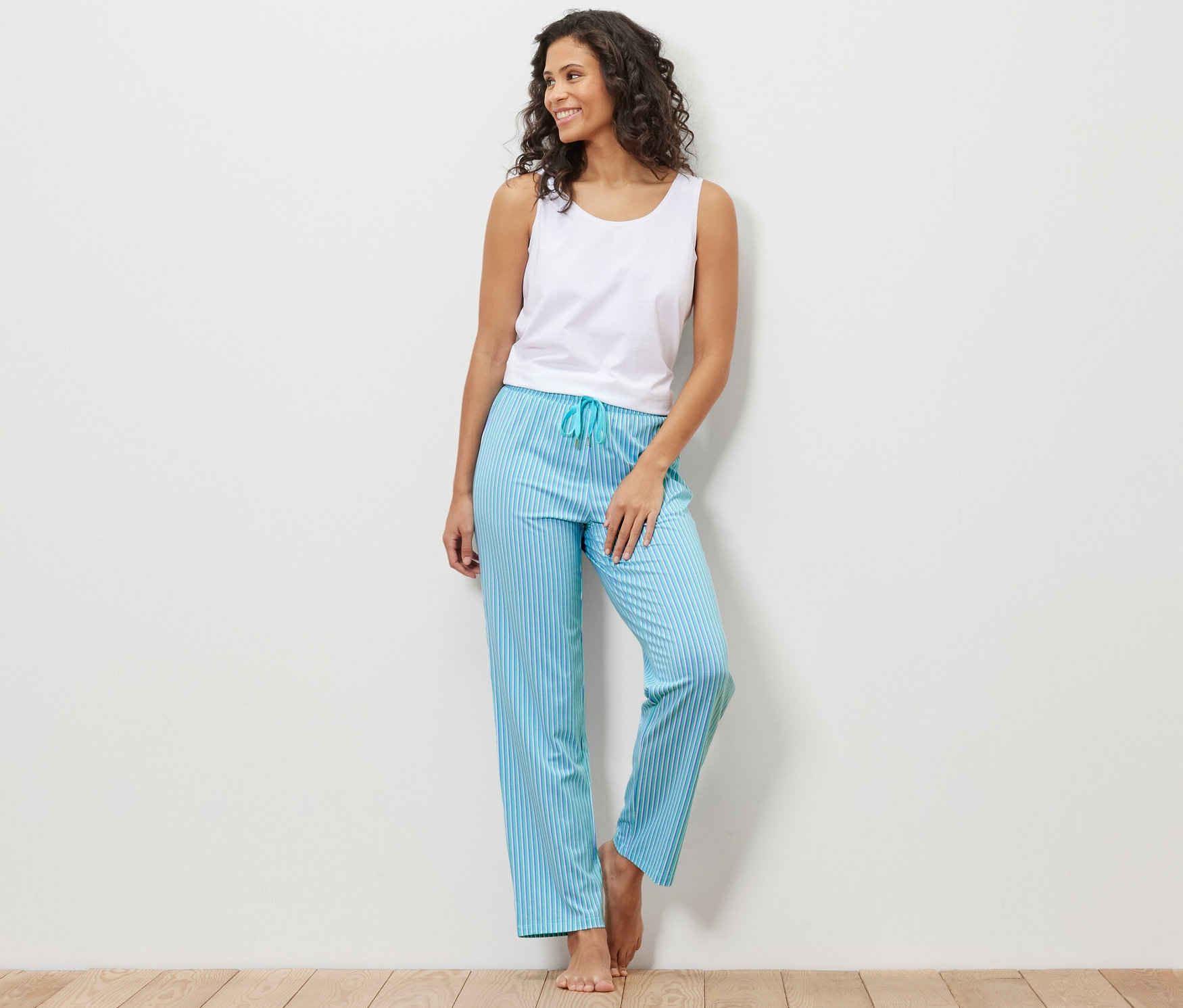 Frau, an eine Wand gelehnt, trägt ein weißes Tanktop und eine blaue, gestreifte Pyjamahose.