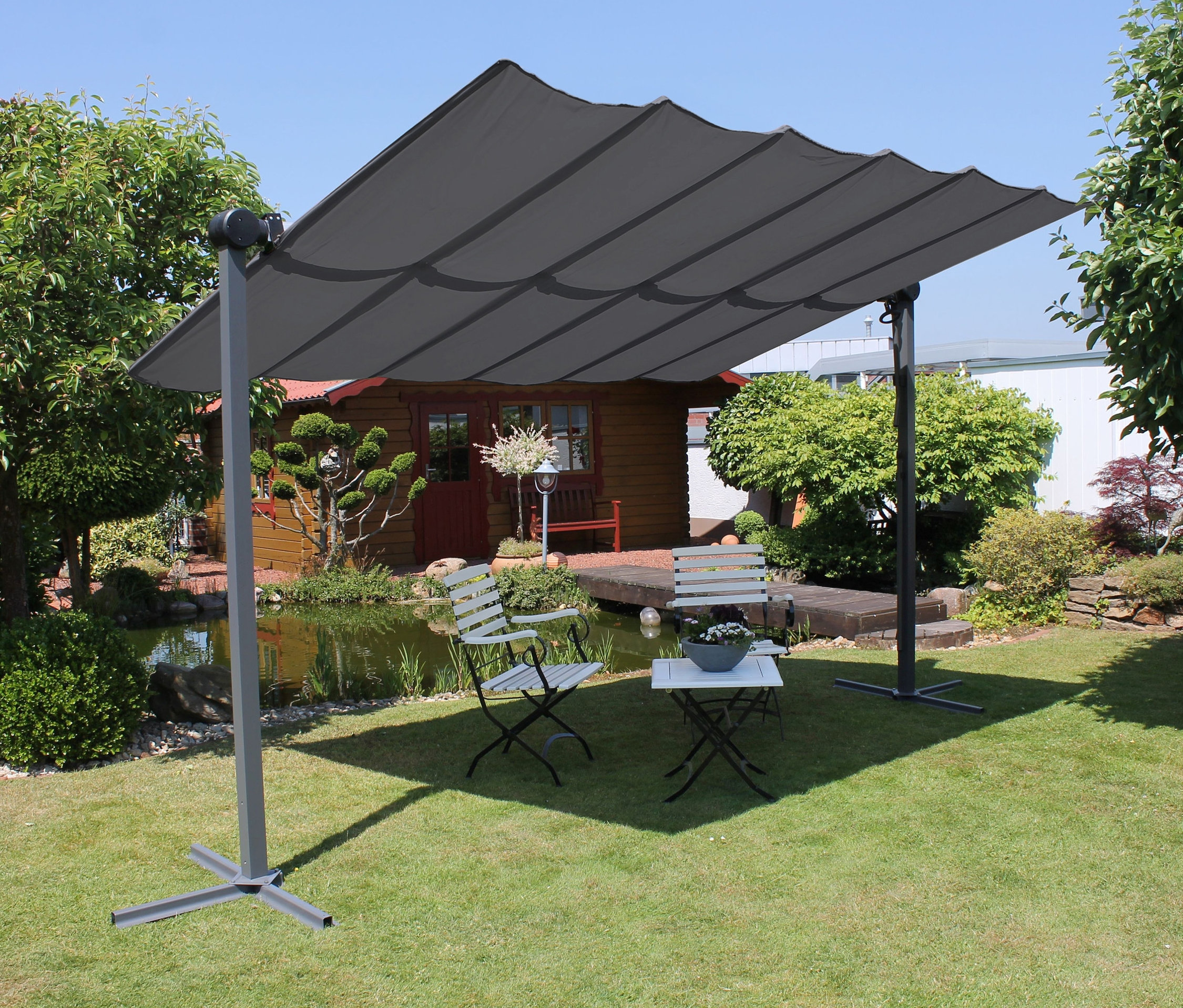 Eine anthrazitfarbene Leco Sonnenmarkise, 400 x 250 cm, steht auf einer Wiese und beschattet weiße Gartenmöbel.