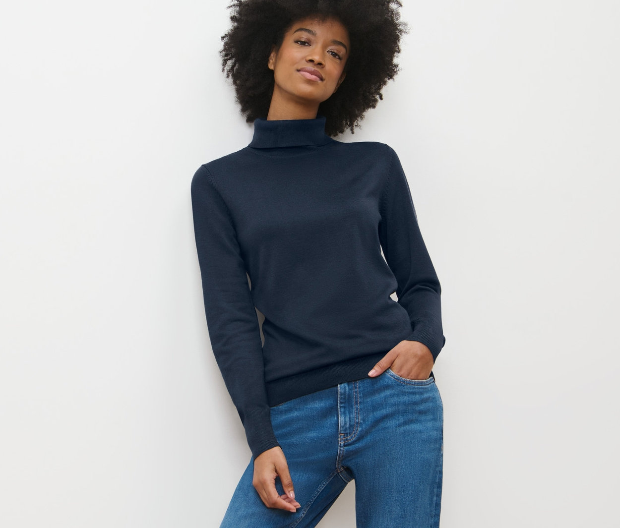 Frau mit Afro trägt dunkelblauen Feinstrick-Rollkragenpullover und Jeans.