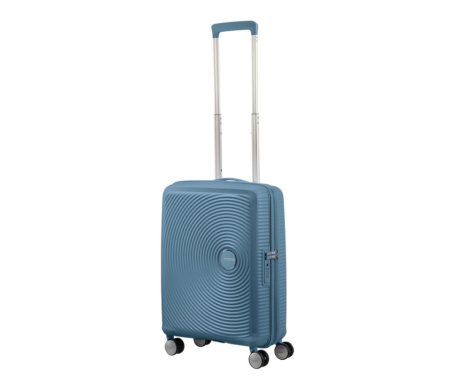 Ein blauer American Tourister Hartschalen-Koffer »Soundbox« Spinner, klein, stone blue auf einem weißen Hintergrund.