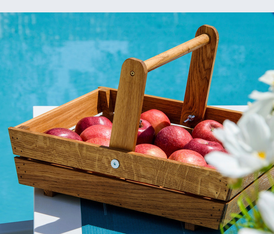 Ein hölzerner Erntekorb gefüllt mit roten Äpfeln steht auf einem Tisch neben einem Pool.