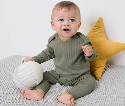 Baby sitzt auf einer grauen Strickdecke neben einem gelben Stern. Es trägt ein 3er-Pack Baby-Rippshirts.