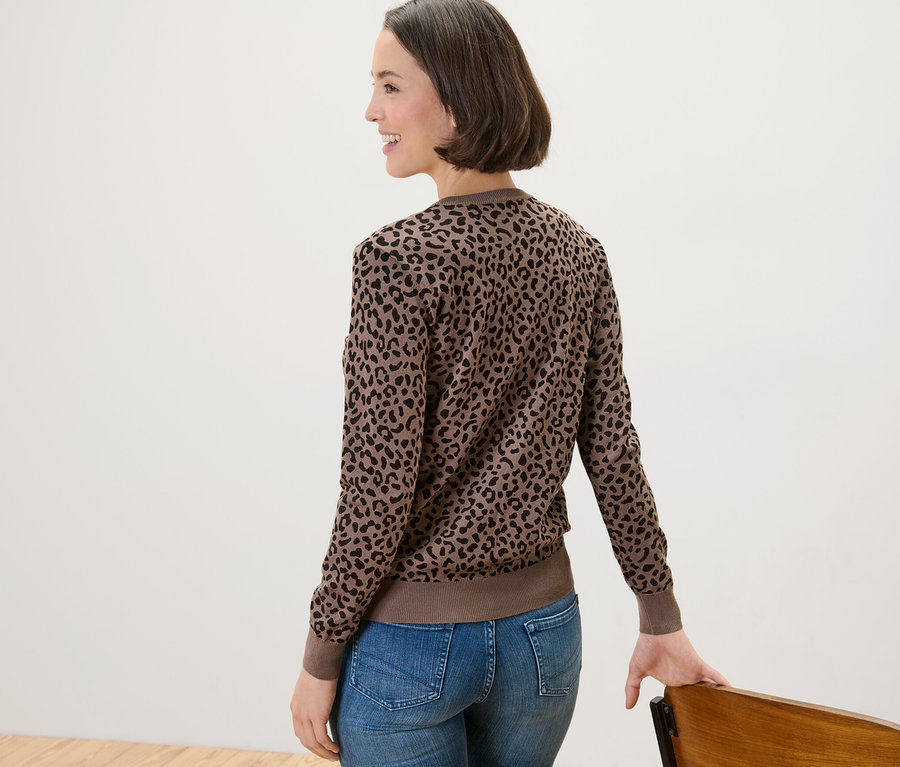 Frau mit Jeans und bedrucktem Feinstrick-Cardigan posiert neben einem Stuhl.