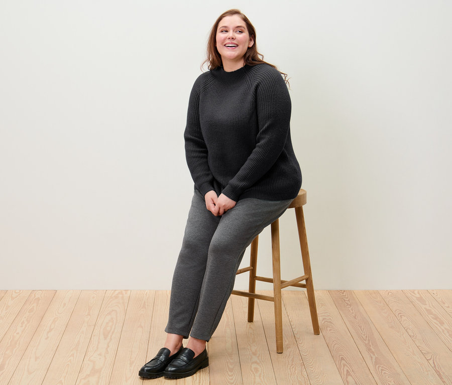 Eine Frau sitzt auf einem Hocker und trägt einen schwarzen Grobstrickpullover mit Stehkragen und eine Joggpants.