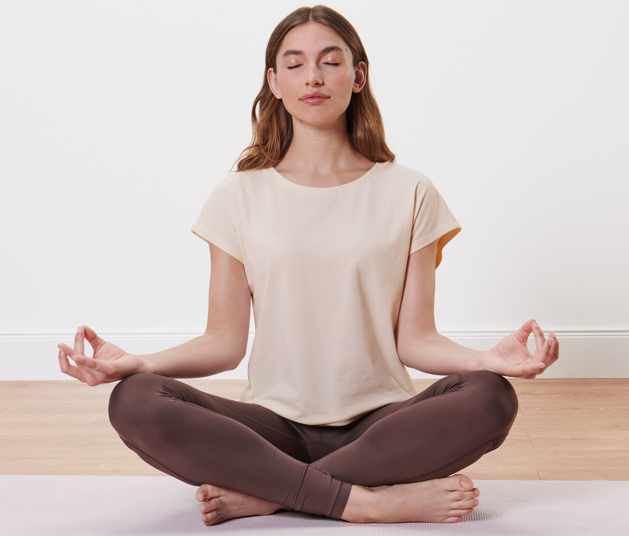 Frau sitzt im Schneidersitz auf einer Matte und trägt ein offwhite Sportshirt.