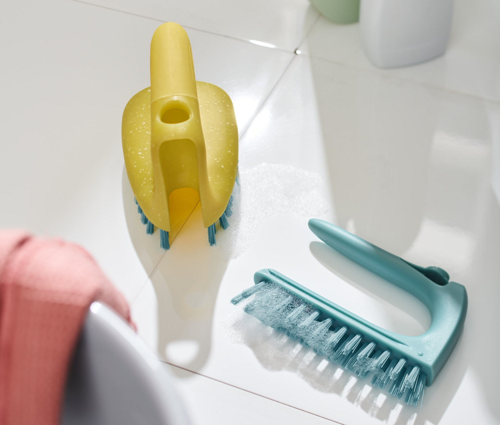 Eine gelbe 2-in-1-Fliesen-Bodenbürste mit blauen Borsten liegt auf einem weißen Boden neben einer blaugrünen Bürste.