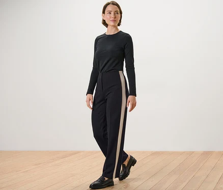 Frau posiert in schwarzem Langarmshirt und schwarzer Jerseyhose mit Galonstreifen.
