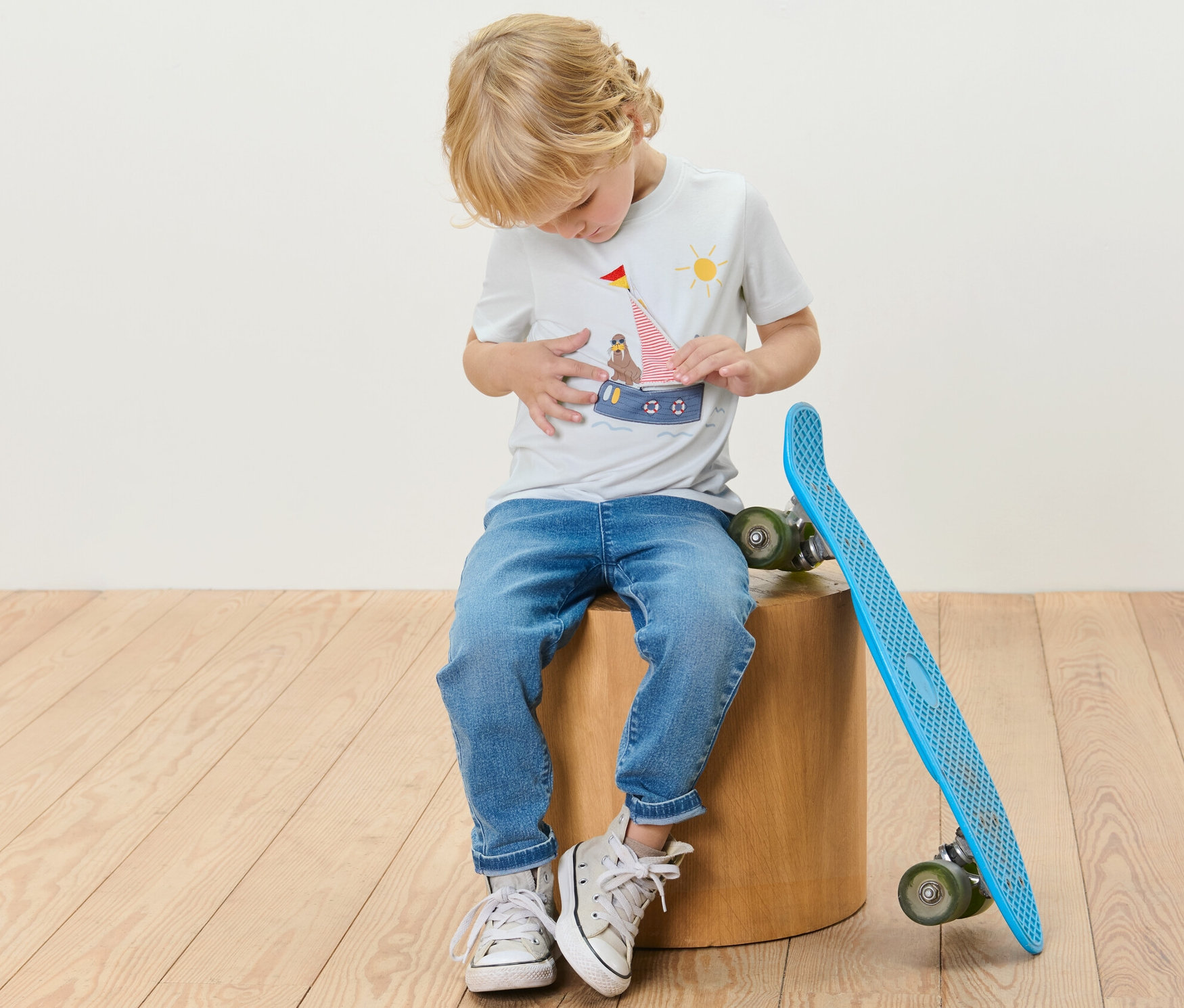 Ein Junge sitzt auf einem Holzschemel neben einem blauen Skateboard und trägt ein T-Shirt mit maritimer Applikation.