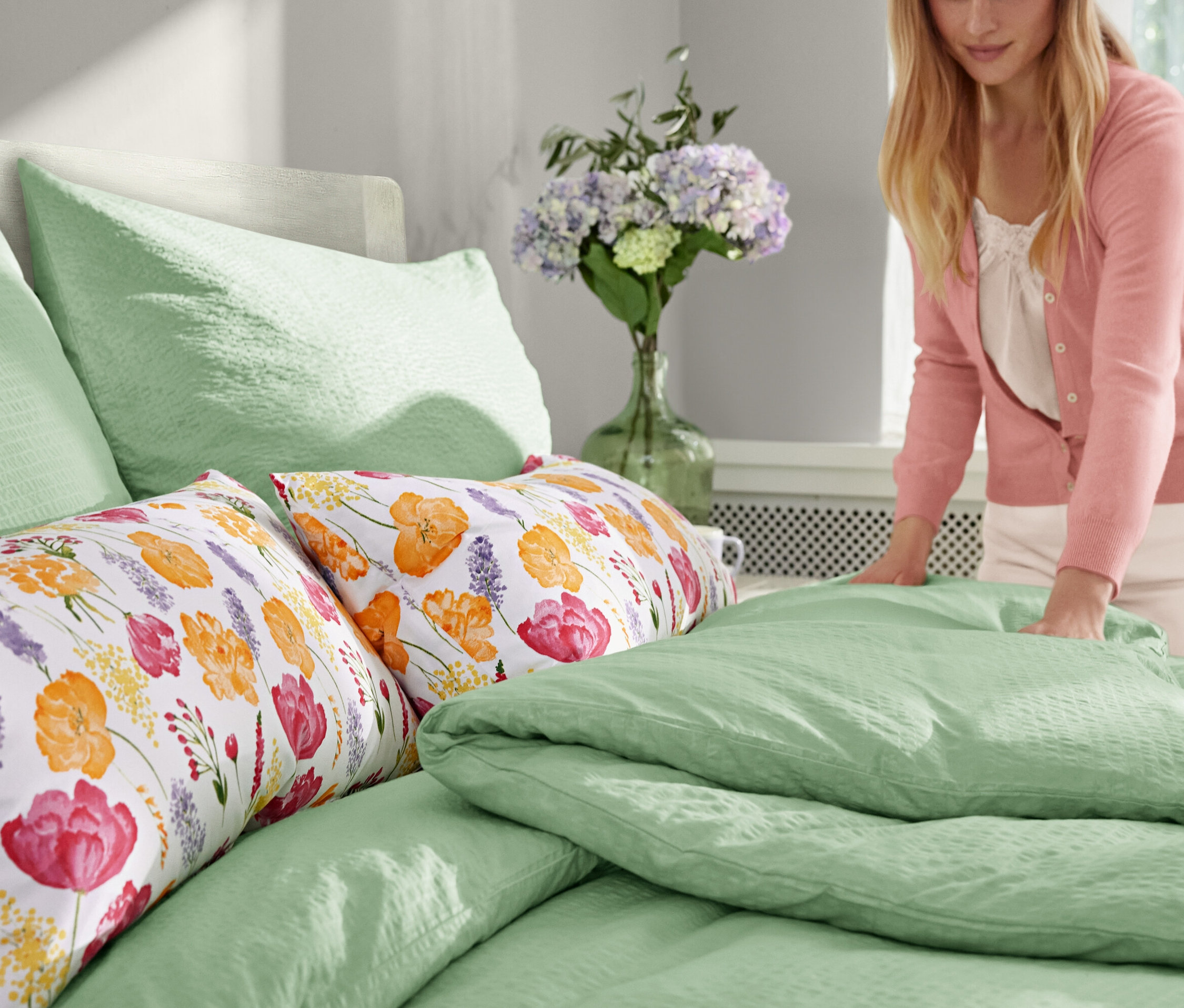 Eine Frau macht ein Bett mit einem hellgrünen Jersey-Spannbettlaken in Normalgröße und Seersucker-Bettwäsche in Normalgröße. Auf dem Bett liegen zwei Microfaser-Kopfkissenbezüge und auf dem Nachttisch stehen zwei LED-Echtwachskerzen im Glas.