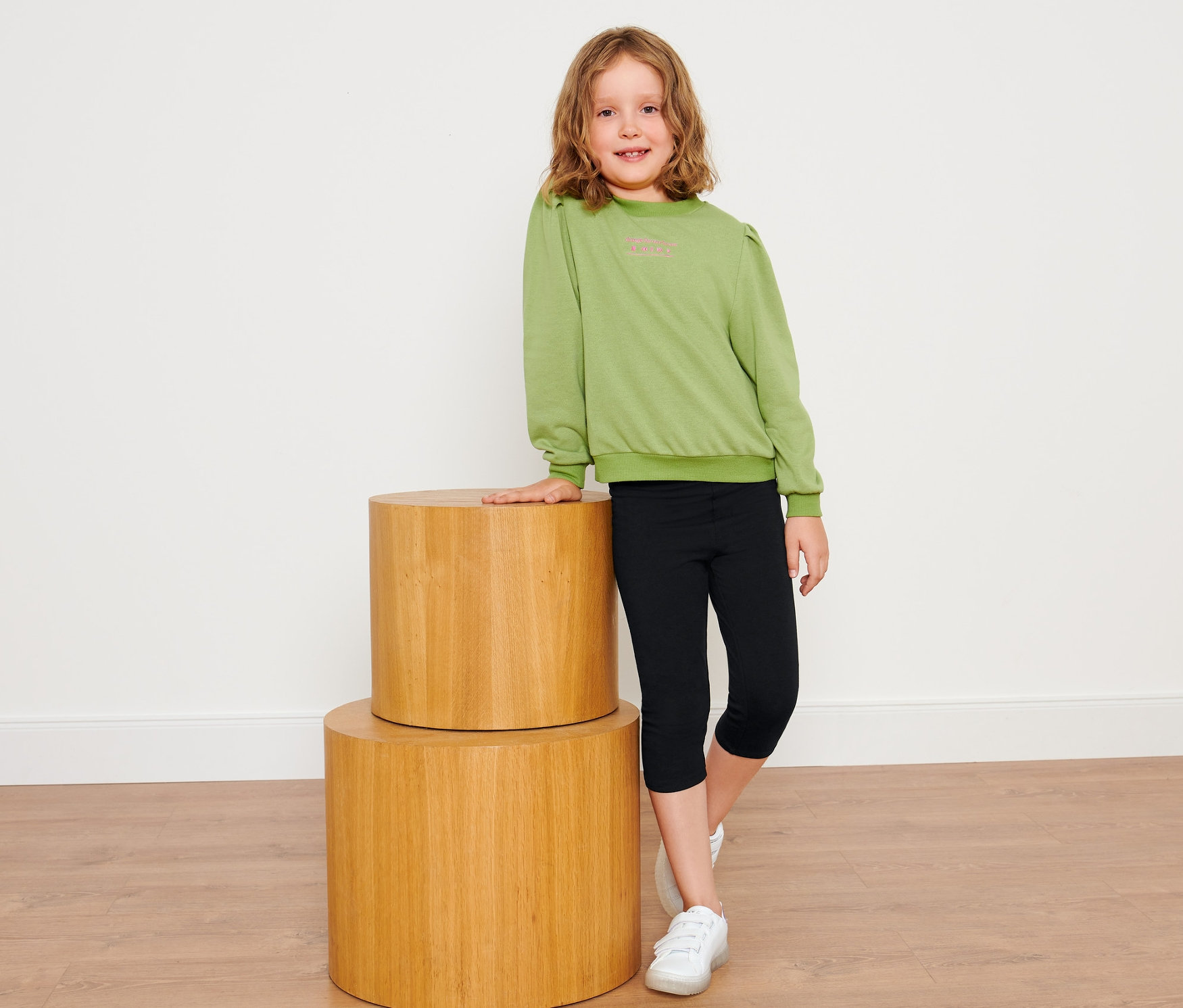 Mädchen posiert neben zwei Holzhockern, trägt einen grünen Pullover und schwarze 3/4-Kinderleggings (2 Stück).