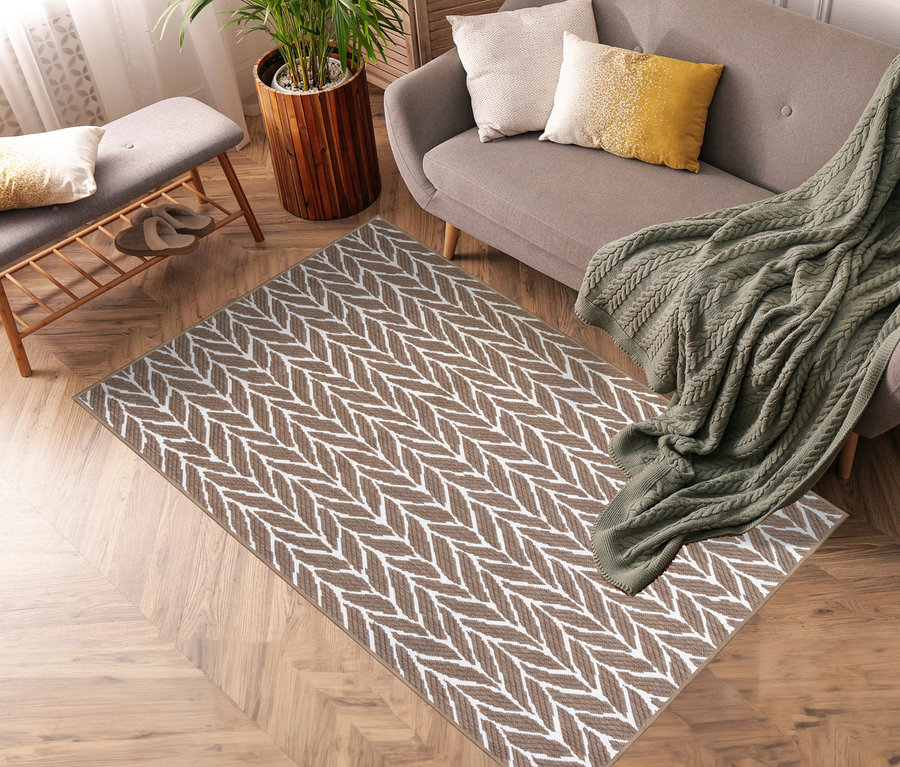 Ein brauner und weißer Outdoor-Wendeteppich »Knitted Joy« liegt auf einem Holzboden in der Nähe eines grauen Sofas mit Kissen und einer grünen Strickdecke.