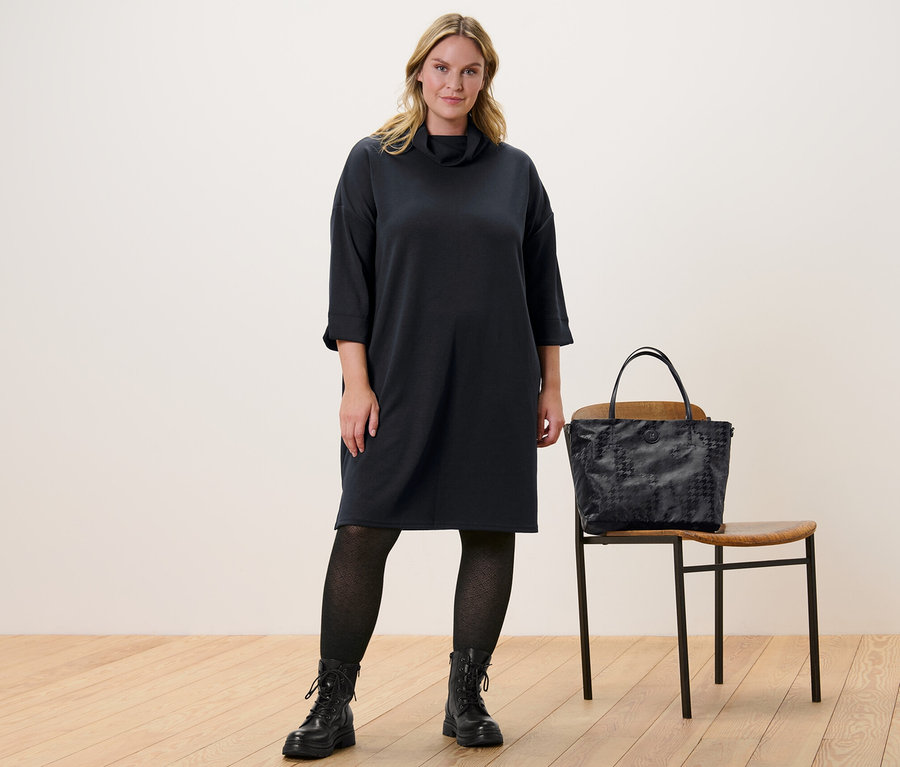 Eine Frau steht und trägt ein Sweatkleid aus Modal-Mix und eine Strickstrumpfhose mit Struktur. Neben ihr steht ein Stuhl, auf dem eine Handtasche liegt.