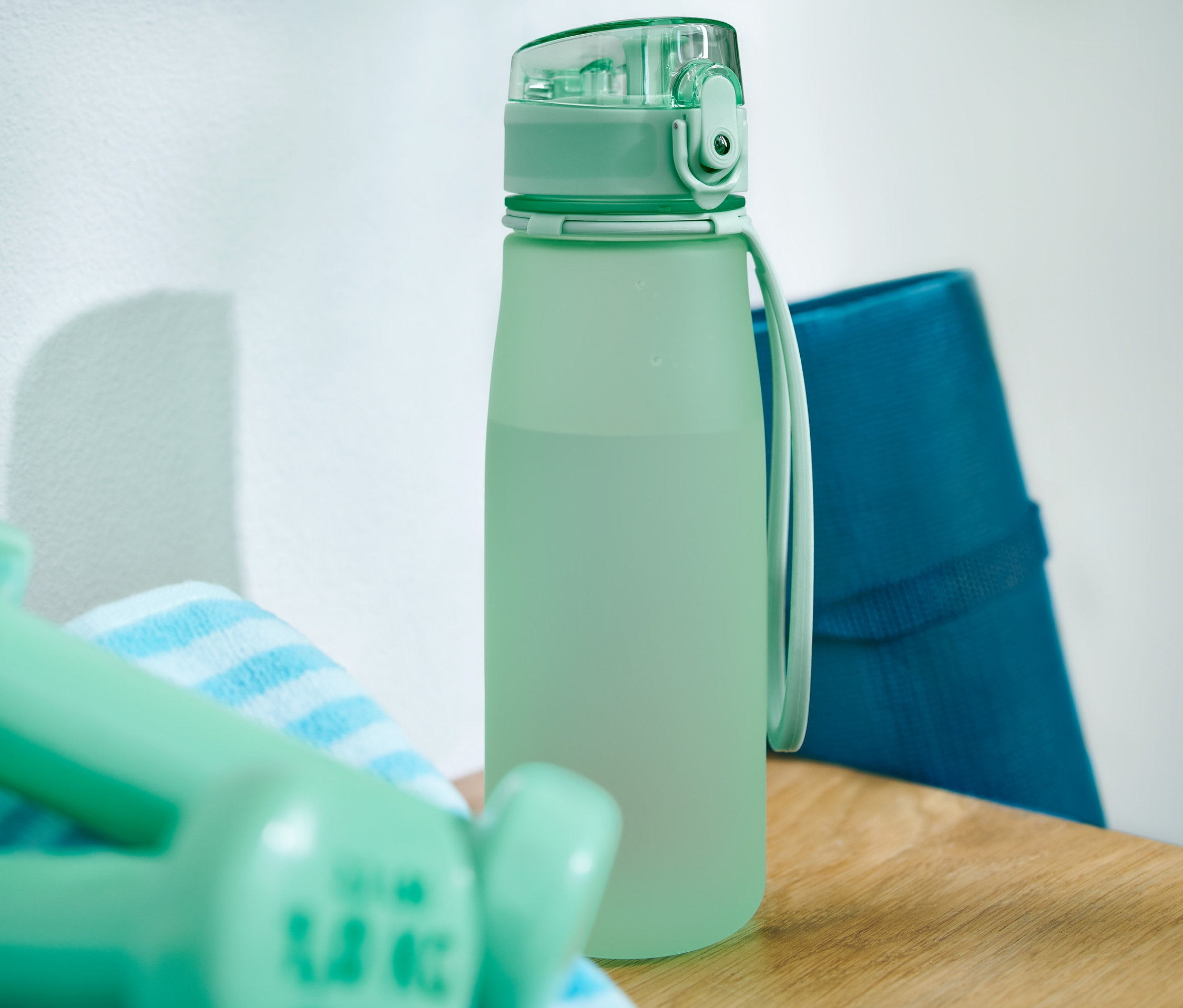 Grüne Sport-Trinkflasche steht auf einem Holztisch neben Handtuch, Hanteln und blauer Übungsmatte.