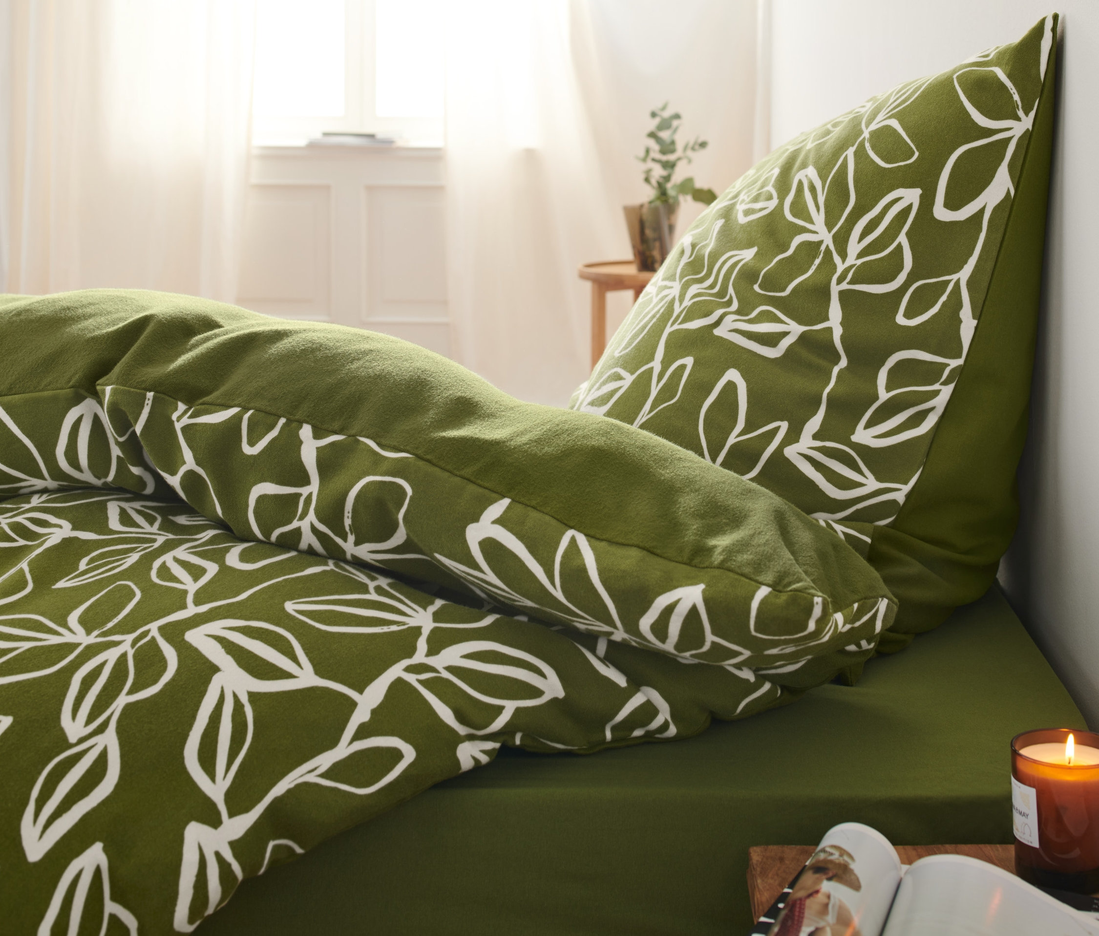Grüne Bettwäsche mit weißem Pflanzenmuster auf einem Bett neben einer brennenden Kerze.