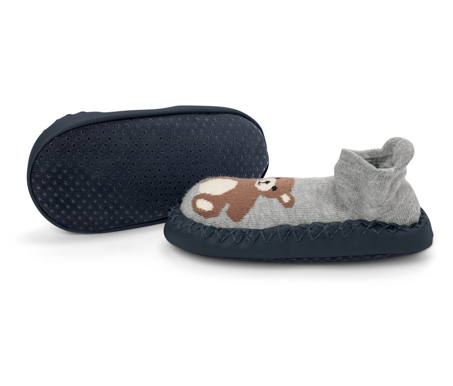 Ein grauer Kinderhausschuh mit Bär liegt neben einer dunkelblauen Anti-Rutsch-Sohle.