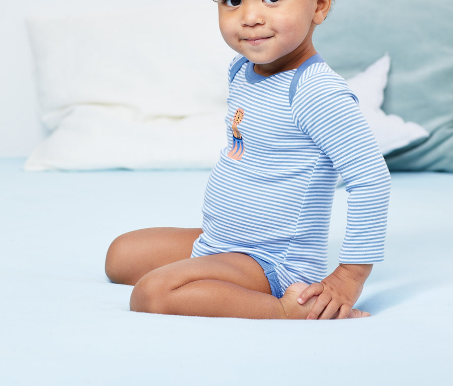 Ein Kind sitzt auf einem blauen Bett und trägt einen blauen, gestreiften Langarmbody. Das Kind trägt einen von '3 Langarm-Bodys'.