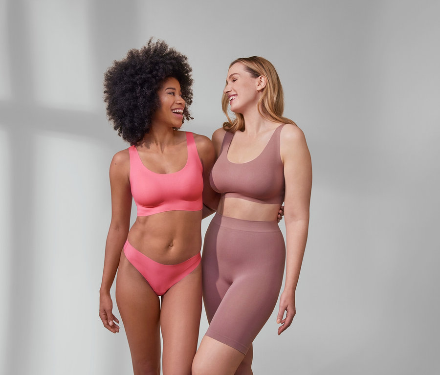 Zwei Frauen umarmen sich lachend. Eine trägt ein pinkes Komfort-Bustier »bonded« und 5 Magic-Cut-Strings. Die andere trägt Seamless-Shorts und ein Oberteil.