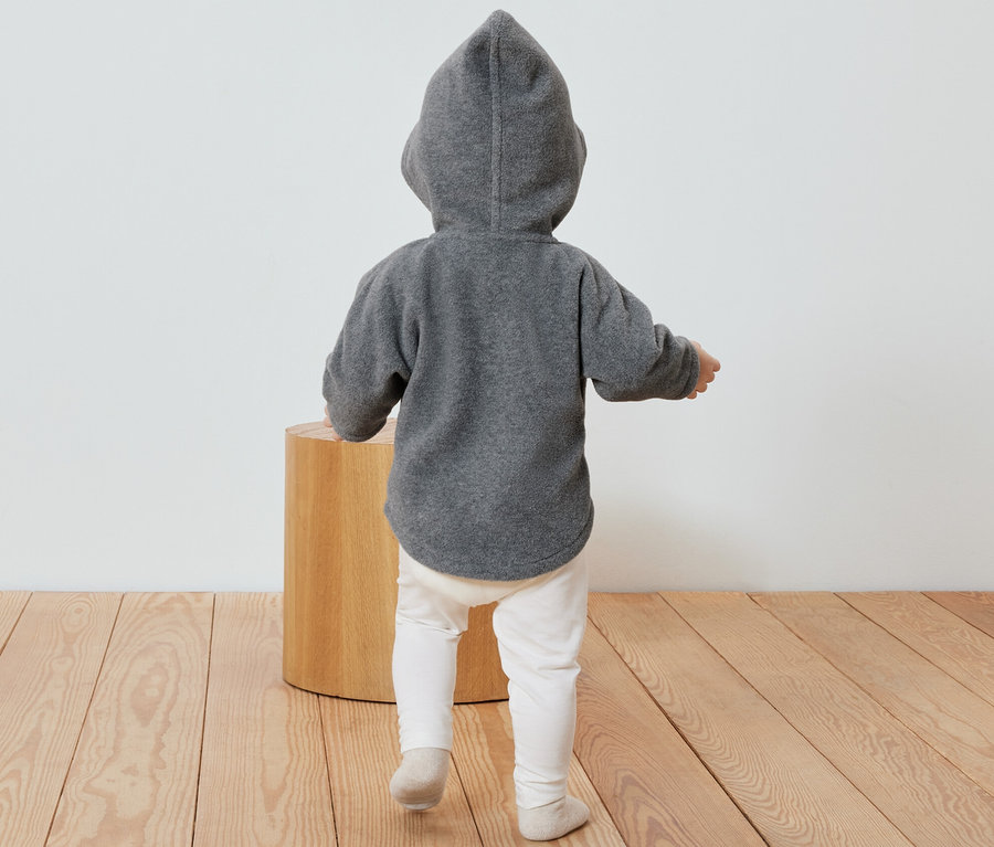 Ein Kind steht mit dem Rücken zur Kamera und trägt eine graue Baby-Fleecejacke mit Kapuze.